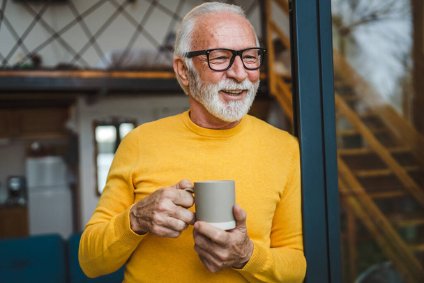One man senior caucasian male with beard and eyeglasses stand on balcony at door in day happy smile hold cup of coffee copy space morning daily routine concept - Photo, Image