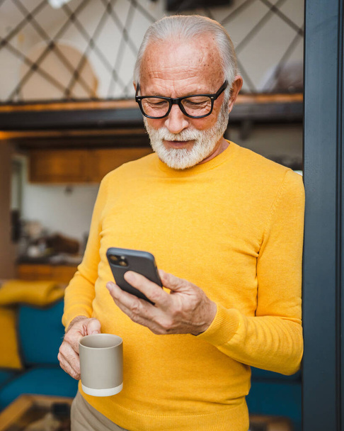 Jeden starszy człowiek ze stojakiem na brodę na balkonie trzymać telefon komórkowy i filiżankę kawy codziennie rano rutyna prawdziwa osoba kopiować przestrzeń czytać lub używać smartfona Internet przeglądać sieci społecznościowe - Zdjęcie, obraz