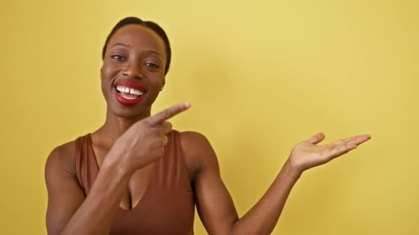 アフリカ系アメリカ人女性は驚きに立ち,明るく微笑み,孤立した黄色の背景にある空間を指差しながら,自信を持って手で提示します.. - 映像、動画