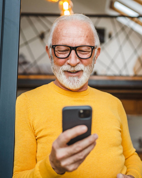 One senior man with a beard stand on the balcony hold a mobile phone and cup of coffee happy smile daily morning routine real person copy space read or use smartphone internet browse social network - Photo, Image