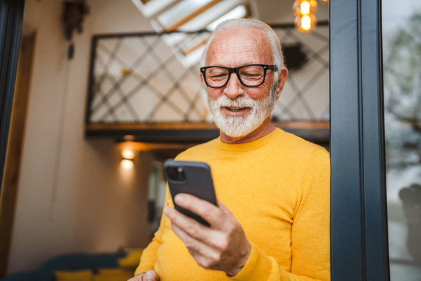 Ein älterer Mann mit Bart steht auf dem Balkon und hält ein Mobiltelefon und eine Tasse Kaffee glücklich lächeln täglichen Morgen Routine echte Person Kopierraum lesen oder Smartphone nutzen Internet-Browse-soziales Netzwerk - Foto, Bild