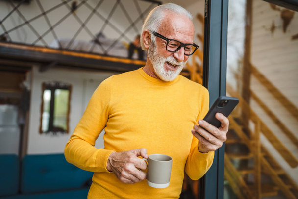 Un hombre mayor con un soporte de barba en el balcón sostiene un teléfono móvil y una taza de café sonrisa feliz rutina diaria de la mañana espacio de copia en persona real leer o usar el teléfono inteligente Internet navegar por la red social - Foto, imagen