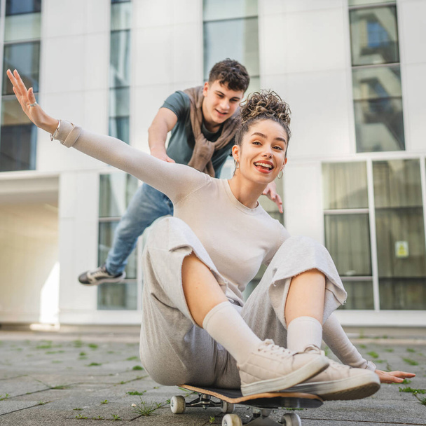 Young man and woman teenager couple caucasian brother and sister have fun on skateboard pushing to ride by the modern building in day happy smile real people family love concept copy space - Photo, Image
