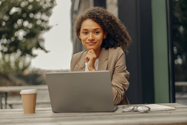 Hymyilevä naispuolinen freelancer työskentelee kannettavan tietokoneen kanssa istuessaan kahvilan terassilla. Etätyön käsite - Valokuva, kuva