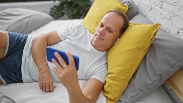 Улыбающийся мужчина среднего возраста, лежащий в постели, бодрствующий, погруженный в просмотр видео на своем смартфоне в своей уютной спальне - Кадры, видео