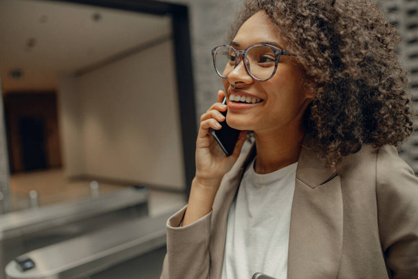 Улыбающаяся деловая женщина разговаривает по телефону с клиентом, стоя на офисном фоне - Фото, изображение