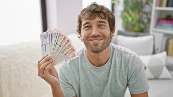 Delighted young man, radiating positivity, confidently holds colombian pesos; his joy reflected in a dazzling, toothy smile at home. - Footage, Video