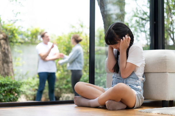 Stressed and unhappy young girl huddle in corner, cover her ears blocking sound of her parent arguing in background. Domestic violence at home and traumatic childhood develop to depression. Synchronos - Photo, Image