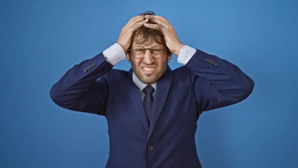Young business man stressed with severe headache, clutching his head in pain. isolated on blue background, bearing an expression of suffering from migraine. - Footage, Video