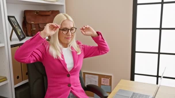 Junge, selbstbewusste blonde Geschäftsfrau, müde und doch lächelnd, streckt ihren Rücken, während sie an ihrem eleganten Bürotisch gekonnt am Laptop arbeitet - Filmmaterial, Video