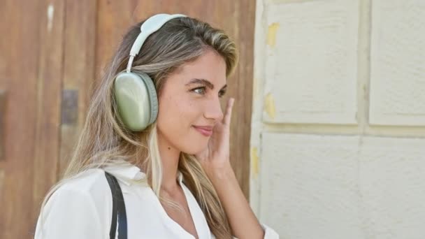 Młoda blondynka słuchająca muzyki uśmiechającej się na ulicy - Materiał filmowy, wideo