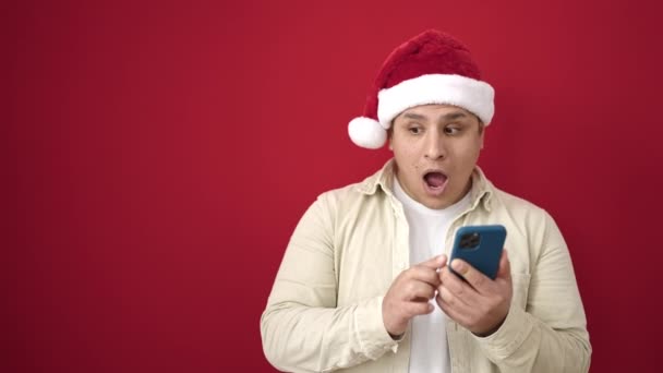 Νεαρός Ισπανός που φοράει χριστουγεννιάτικο καπέλο χρησιμοποιώντας smartphone σε απομονωμένο κόκκινο φόντο - Πλάνα, βίντεο