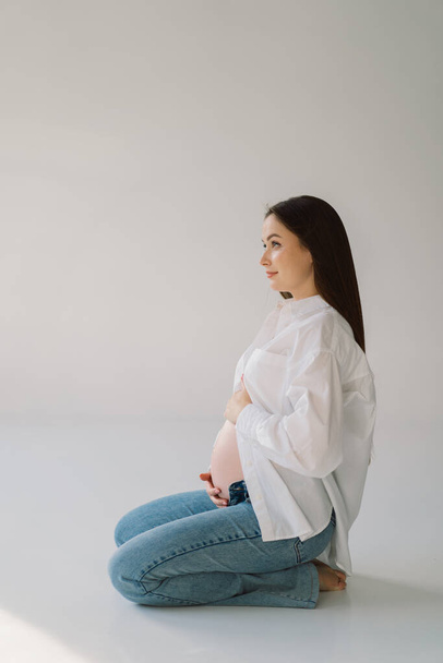 Linda joven embarazada sentada en el estudio blanco en luz cálida con las manos en el estómago embarazada en el fondo de la pared con sombra. Concepto de embarazada, espacio libre, disfrutando del embarazo. Cuerpo embarazado. - Foto, Imagen