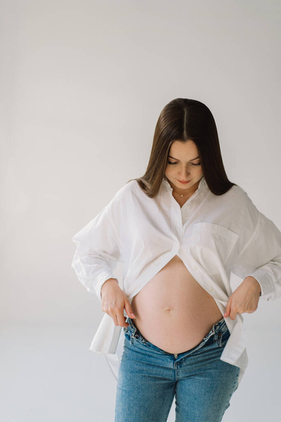 Schattig jong zwanger meisje zitten op de witte studio in warm licht met de handen op zwangere maag op de achtergrond van de muur met schaduw. Zwanger concept, vrije ruimte, genieten van de zwangerschap. Zwanger lichaam. - Foto, afbeelding