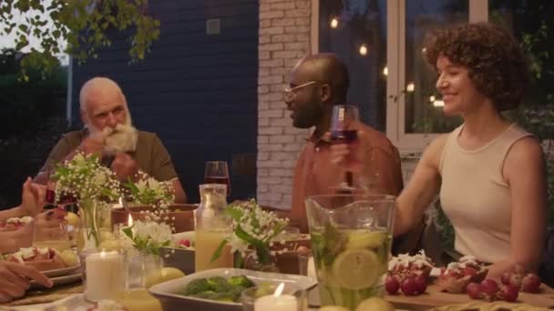Grupo de diversos miembros de la familia sentados en la mesa festiva al aire libre en la noche de verano cenando y hablando - Metraje, vídeo