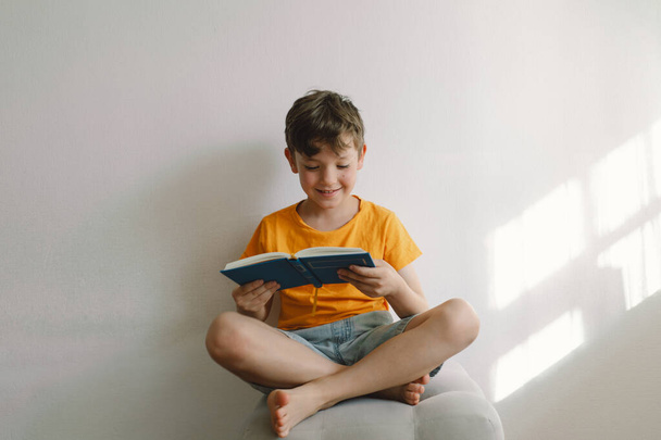 Ein süßer Junge in einem orangefarbenen T-Shirt sitzt auf einem weichen Hocker und liest ein Buch. Kopierraum. Echte Menschen und Lebensstil - Foto, Bild