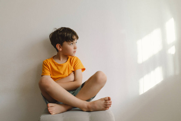 Cute boy sitting barefoot in a home. Beautiful light. Brunette boy 7 years old, casual clothes. Cozy home and lifestyle. Indoors. - Photo, Image