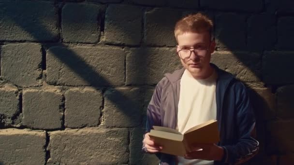 Mittleres Porträt eines jungen kaukasischen Mannes mit Brille, der vor einer dunklen Betonmauer steht und Buch liest und dann in die Kamera blickt - Filmmaterial, Video