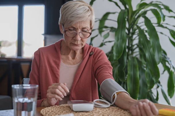 Senior vrouw zit alleen thuis, meten van haar bloeddruk met een huishoudapparaat - Foto, afbeelding