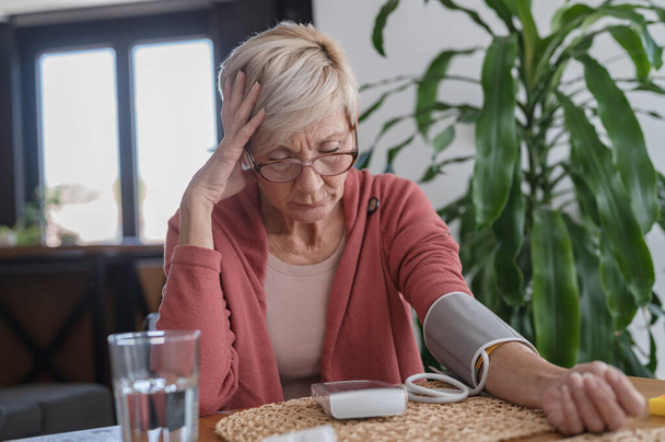 Seniorin sitzt allein zu Hause und misst ihren Blutdruck mit einem Haushaltsgerät - Foto, Bild