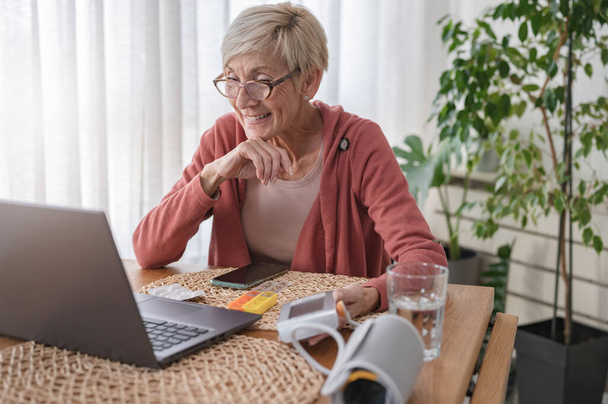 Uma mulher idosa sentada na frente de um laptop em casa, tendo uma chamada de vídeo on-line com um médico. Consulta médica online - Foto, Imagem