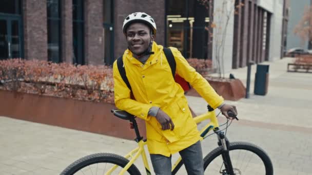 Mediano tiro largo de mensajero masculino mirando a la cámara y sonriendo, tiene equipo de entrega de alimentos, casco, chaqueta amarilla, mochila y bicicleta - Metraje, vídeo