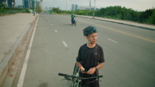 Tilt down shot of cyclist in sportswear and helmet standing with bike on road, using navigation app on smartphone and looking around - Metraje, vídeo