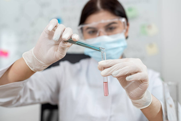 Kobieta kaukaska asystentka laboratoryjna wlewa próbkę do kolby w laboratorium. Eksperyment z odczynnikami mieszającymi w probówce  - Zdjęcie, obraz