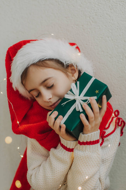 A cute teenage girl in a pullover and Santa Claus hat holding a box with a gift against the background of a light wall. Magic Christmas atmosphere at home. - Photo, Image