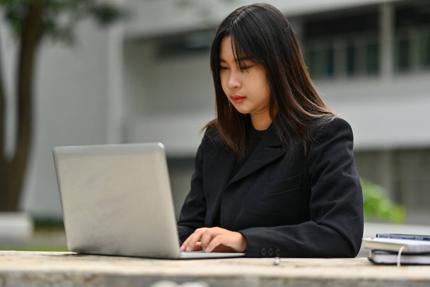 Ασιάτισσα επιχειρηματίας που εργάζεται με φορητό υπολογιστή ενώ κάθεται έξω από το κτίριο. - Φωτογραφία, εικόνα