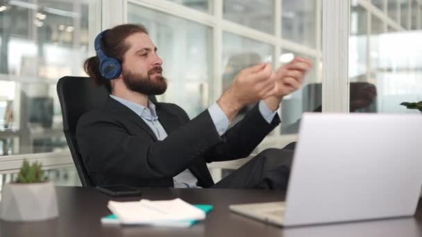Smiling caucasian businessman wearing wireless headset imagining himself as orchestra conductor while waving virtual baton. Active male melomaniac listening classic music and putting feet on table. - Footage, Video