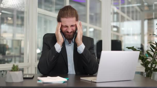 Frustrovaný vyčerpaný obchodník, který se dotýká hlavy, protože cítí bolest, když sedí u stolu před přenosným laptopem. Rozrušit unavený bělošský zaměstnanec pomocí moderní gadget pro práci v kanceláři. - Záběry, video