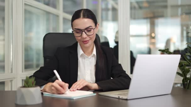 Optimista sikeres nő ül a munkahelyen és információt ír a digitális számítógépről a notebook az irodában. Fókuszált elegáns barna zárt sötét kabát mosolyog és jegyzetel beltéri. - Felvétel, videó