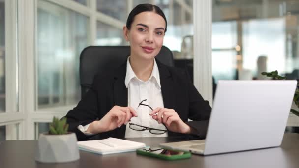 Mujer emprendedora sonriente vestida con ropa de negocios negra sentada en una silla de oficina y usando dispositivos digitales mientras sostiene las gafas en las manos en una habitación espaciosa. Concepto de éxito y confianza. - Metraje, vídeo