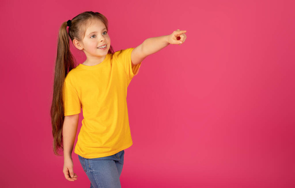 Koukni na tohle. Roztomilé holčička ukazující pryč na kopírovat prostor s prstem při stání nad růžové studio pozadí, usmívající se předpuberťačka dítě demonstrující volné místo pro design nebo nabídku - Fotografie, Obrázek
