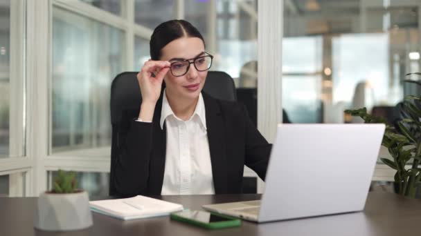 Beautiful successful female sitting in front of wireless devices and adjusting glasses while smiling in office. Cheerful lady dressed in classic style showing harmony and career achievements indoors. - Footage, Video