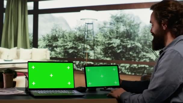 Imprenditore lavora con schermo verde su laptop e tablet, seduto alla scrivania nella sua elegante cabina alpina moderna. Ricco uomo d'affari che esamina display mockup isolati su due gadget, miliardario. - Filmati, video