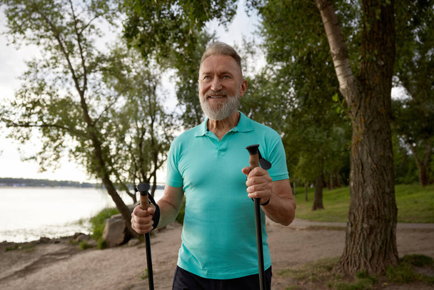 Портрет пожилого человека, наслаждающегося красивыми пейзажами парка во время похода летом. Взрослый спортсмен в возрасте, занимающийся спортивной ходьбой на шесте для улучшения физической выносливости и здравоохранения - Фото, изображение