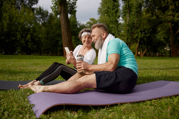 Seniorenpaar zittend op fitnessmatten rusten na yoga training buiten in stadspark. Oudere vrouw die smartphone gebruikt, volwassen man die water drinkt. Sportweekend en vrije tijd bij pensionering - Foto, afbeelding