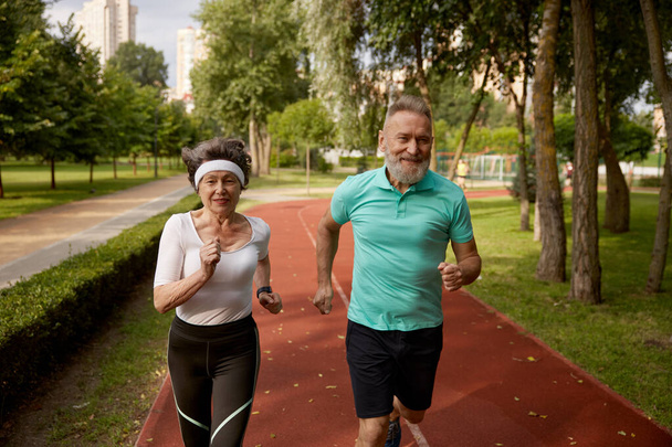 Senior couple running on track together at natural park. Happy healthy elderly woman and mature man friends exercising outdoors - Photo, Image