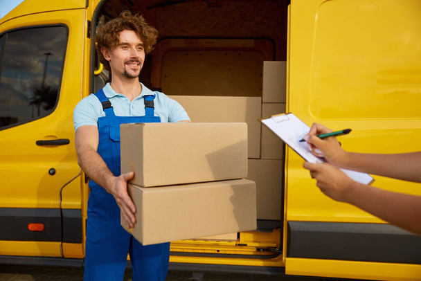 Friendly smiling young courier giving clipboard with checklist and pen for signature. Deliveryman standing nearby minivan full of cardboard packages. Logistics worker portrait - Photo, Image