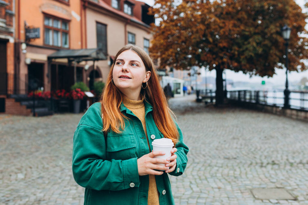 Счастливая рыжая женщина в модной одежде держит чашку эко чая на фоне старого города. Модель, стильная девушка, гуляющая на свежем воздухе, держит чашку кофе на вынос. - Фото, изображение