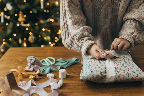 Упаковка рождественских подарков. Руки в уютном свитере, обертывающем стильный подарок в праздничную оберточную бумагу с лентами, винтажными украшениями, бантами на деревянном столе. Атмосферный зимний отдых - Фото, изображение