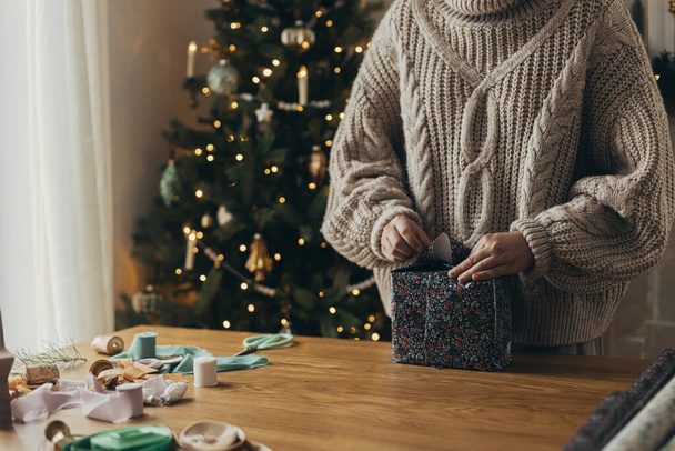 Mujer en suéter acogedor envolviendo elegante regalo de Navidad en papel de regalo festivo con cintas, adornos vintage, arcos en la mesa de madera. Vacaciones de invierno atmosféricas, envolviendo regalo de Navidad - Foto, Imagen