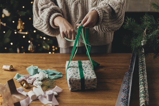 Обгортання різдвяних подарунків. Руки в затишному светрі обгортають стильним подарунком у святковому обгортковому папері стрічками, вінтажними прикрасами, бантами на дерев'яному столі. Атмосферні зимові свята - Фото, зображення