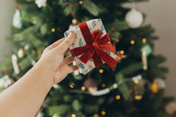 Hand houden stijlvolle kerstcadeau in feestelijke verpakking papier met strik op de achtergrond van kerstboom verlichting. Fijne feestdagen. Sfeervolle kersttijd - Foto, afbeelding