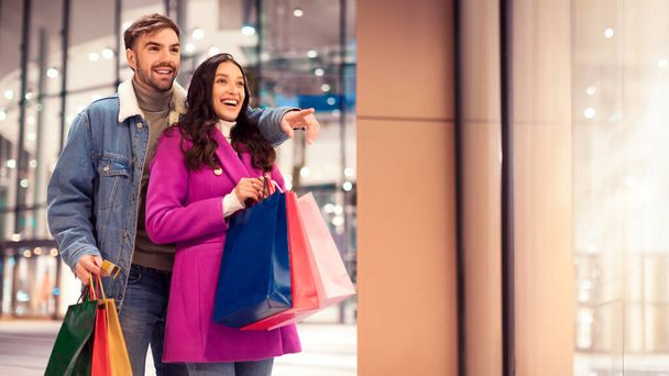 Happy young couple pointing and showing something exciting standing with shopping bags, outside of store on winter evening, perfect for seasonal sale ads, city mall night scene with free space - Photo, Image