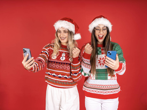 Fiatal női barátok viselnek karácsonyi kötött pulóver Mikulás kalap hold használata mobiltelefon do győztes gesztus összeszorított öklök piros háttér. Boldog új évet ünneplés téli ünnep koncepció, másolás tér. - Fotó, kép