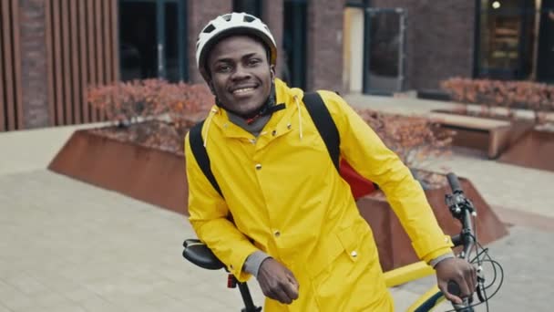 Porträt eines afroamerikanischen Zustellers, der lächelnd für die Kamera posiert, während er mit dem Fahrrad auf der Straße der Stadt steht - Filmmaterial, Video
