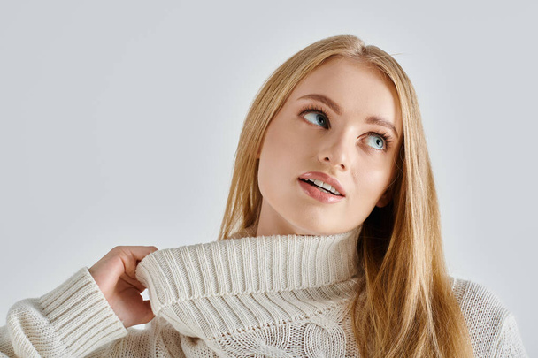charming and dreamy woman stretching collar of white knitted sweater and looking away on grey - Photo, Image